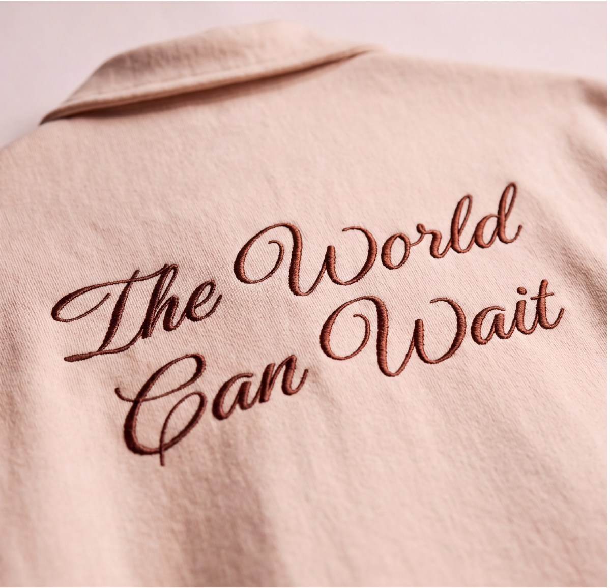 A close-up of an oversized Boxy fit shirt. The front features a matching embroidered geometric logo on the left chest and the word 'The World Can Wait' in embroidery at the back.