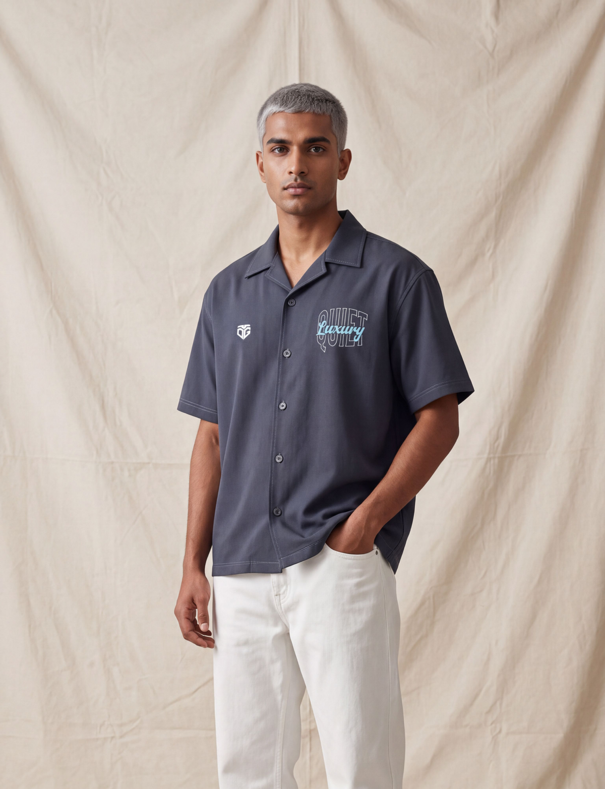 A man stands against a beige fabric backdrop wearing a dark grey boxy fit shirt style 'Quite Luxury' graphic on the chest, paired with off-white trousers and dark sneakers.